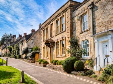 Reihe traditioneller honigfarbener Steinhäuser entlang einer Straße in Burford mit gepflegten Gärten und Kirchturm im Hintergrund.
