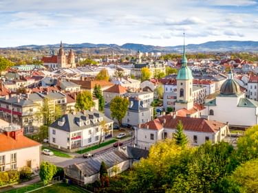 Luftaufnahme von Nowy Sącz mit bunten Dächern, weißer Kirche mit grüner Kuppel, historischen Gebäuden und Bergen im Hintergrund.