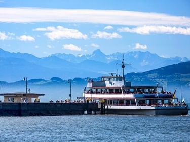 White passenger ferry docked at a pier in Langenargen harbor on Lake Constance, with Alpine mountains and blue sky in the background.