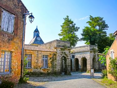 Kopfsteinpflasterplatz in Blaye mit verwitterten Steingebäuden, Torbogen und Kirchturm im Hintergrund. Bäume rahmen die Szene unter blauem Himmel.