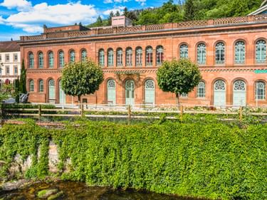 Rotes Backsteingebäude Palais Thermal mit Bogenfenstern in Bad Wildbad. Grüner Efeu bedeckt die Ufermauer, Bäume säumen die Fassade unter blauem Himmel.