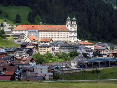 Kloster Disentis mit Zwillingstürmen und orangefarbenem Dach überragt ein Schweizer Alpendorf. Traditionelle Häuser verteilen sich darunter, umgeben von grünen Wiesen und Wald.