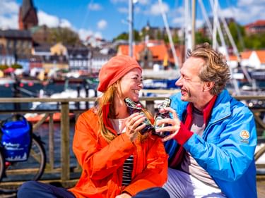 Zwei Radfahrer in bunten Jacken genießen Eis am Flensburger Hafen. Fahrräder und Boote im Hintergrund unter blauem Himmel.