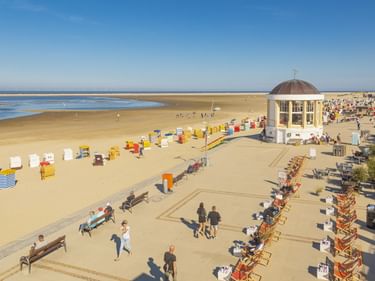 Weißer Musikpavillon mit Kuppel auf der Strandpromenade von Borkum. Bunte Strandkörbe säumen den Sandstrand, mit Reihen orangefarbener Stühle.