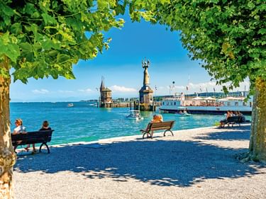 Blick durch die von Bäumen gesäumte Promenade auf den Konstanzer Hafen am Bodensee mit Imperia-Statue, Leuchtturm und Passagierschiffen.
