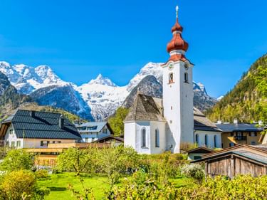 Weiße Kirche mit rotem Zwiebelturm in Lofer, umgeben von traditionellen Alpenhäusern und grünen Wiesen mit schneebedeckten Bergen.