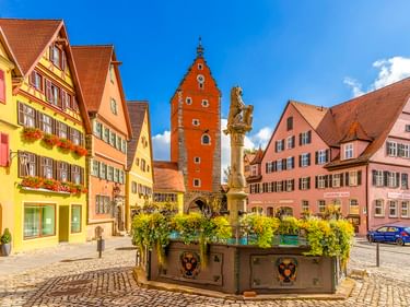 Historischer Marktplatz in Dinkelsbühl mit bunten Fachwerkhäusern, dem roten Wörnitztor, einem Brunnen mit Statue und Kopfsteinpflaster.