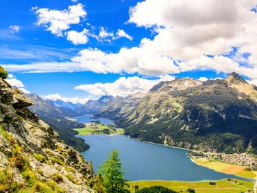 Panoramablick auf den Silsersee mit dem Dorf Silvaplana am Ufer, umgeben von Bergen und grünen Wiesen unter blauem Himmel mit weißen Wolken.