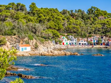 Bucht Cala S'Alguer mit bunten Häusern entlang felsiger Küste. Blaues Mittelmeer, grüner bewaldeter Hang und klarer Himmel sichtbar.