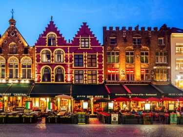 Beleuchtete historische Giebelhäuser am Brügger Grote Markt in der Abenddämmerung mit Außenrestaurants und Kopfsteinpflaster im Vordergrund.