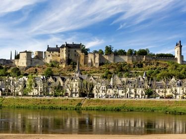 Mittelalterliche Burg auf Hügel über Stadt am Fluss mit traditionellen französischen Gebäuden. Die Loire spiegelt die Szenerie unter blauem Himmel.