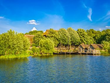 Rekonstruiertes Slawendorf mit strohgedeckten Holzhäusern auf Stelzen am See in Oldenburg, Holstein. Grüne Bäume rahmen die Ufersiedlung.