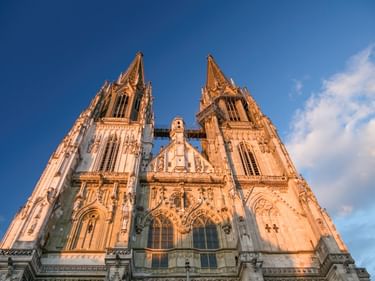 Fassade des Regensburger Doms mit zwei hohen gotischen Türmen vor blauem Himmel. Goldenes Sonnenlicht beleuchtet die Steinarchitektur.