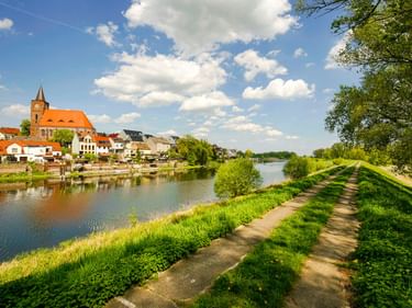 Paved path along a river in Eisenhüttenstadt with church and colorful houses. Green grass banks and trees frame the sunny scene under blue sky.