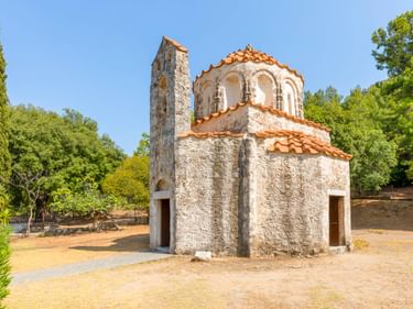 Kleine byzantinische Steinkapelle mit Terrakottadach und Glockenturm auf Rhodos. Die Fountoukli-Kapelle steht auf einer Lichtung umgeben von grünen Bäumen.