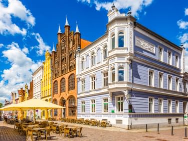 Bunte historische Gebäude am Alten Markt in Stralsund mit gotischer Backsteinarchitektur und Café-Bestuhlung unter gelben Sonnenschirmen.
