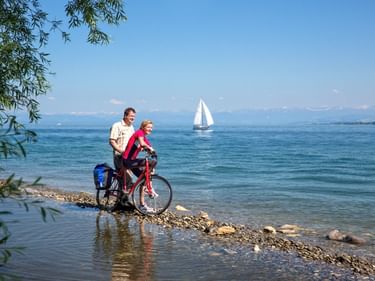 Zwei Radfahrer mit beladenem Fahrrad am steinigen Ufer des Bodensees. Weißes Segelschiff auf blauem Wasser, Berge im Hintergrund, Weidenzweige rahmen die Szene.
