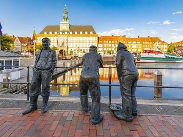 Drei Bronzestatuen von Hafenarbeitern am Emder Hafen mit dem historischen Rathaus und bunten Gebäuden, die sich im Wasser spiegeln.