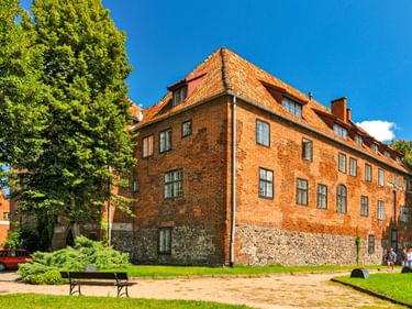 Historische Backsteinburg in Kętrzyn mit rotem Ziegeldach und Steinfundament. Grüne Rasenfläche mit Bänken davor, umgeben von Bäumen.