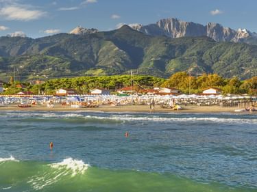 Strand in Forte dei Marmi mit Wellen, Badegästen, weißen Sonnenschirmen und den dramatischen Apuanischen Alpen im Hintergrund unter blauem Himmel.
