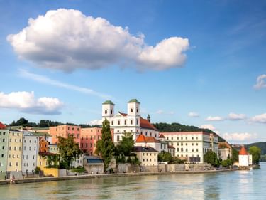 Bunte Häuser am Flussufer in Passau entlang der Donau mit weißer Kirche mit zwei Türmen. Blauer Himmel mit weißen Wolken darüber.