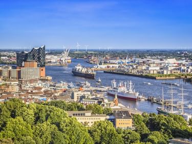 Luftaufnahme des Hamburger Hafens mit Frachtschiffen, Kränen und der Elbphilharmonie. Grüne Bäume im Vordergrund, blauer Himmel darüber.