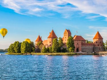 Trakai Island Castle with red brick towers on a lake in Lithuania. Yellow hot air balloons float in the blue sky above green trees.