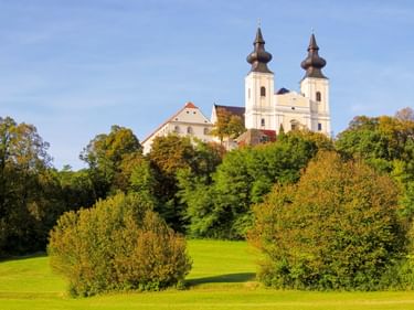 Maria Taferl Basilica with twin onion-domed towers on a green hill in Austria, surrounded by trees and meadows under a blue sky.