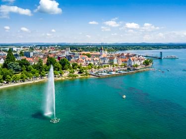 Luftaufnahme von Friedrichshafen am Bodensee mit hoher Wasserfontäne im Vordergrund, historischer Altstadt, Kirchturm und Passagierschiff.