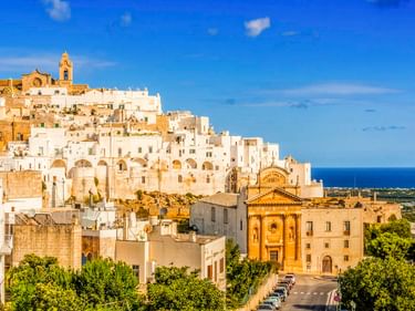 Panoramablick auf Ostuni, eine weiße Hügelstadt in Italien mit historischen Kirchen und Gebäuden, umgeben von grüner Landschaft und blauem Meer.
