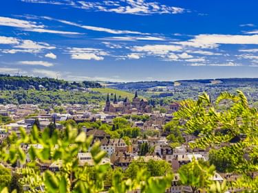 Panoramablick auf Trier mit Domtürmen zwischen Wohngebäuden, eingerahmt von grünem Laub. Hügelige Landschaft unter blauem Himmel.