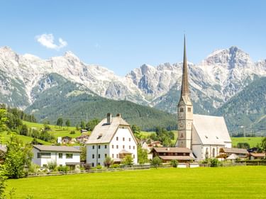 Wallfahrtskirche Maria Alm mit hohem Turm im österreichischen Dorf, umgeben von grünen Wiesen und dramatischen Berggipfeln unter blauem Himmel.