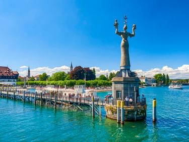 Imperia-Statue auf Sockel im Konstanzer Hafen mit Drehplattform. Blaues Bodenseewasser, Anlegesteg und historische Gebäude sichtbar.