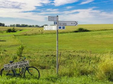 Wegweiser zu Vitt Hafen und Kap Arkona an einem Radweg durch grüne Felder unter blauem Himmel. Ein dekoratives Fahrrad steht neben dem Schild.