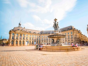Place de la Bourse in Bordeaux mit prächtigem Brunnen im Vordergrund und klassischen Gebäuden aus dem 18. Jahrhundert unter blauem Himmel.