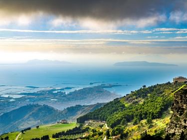 Panoramablick vom Monte Erice mit grünen Hängen, der Küstenstadt Trapani und dem Mittelmeer unter dramatischem Wolkenhimmel.