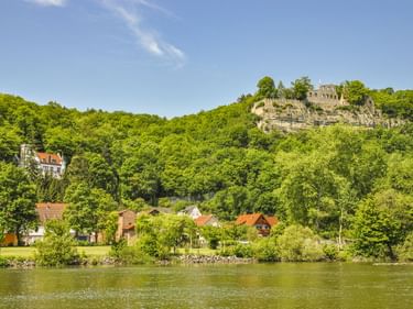 View of Karlsburg castle ruins on a hilltop above Karlstadt with houses along the Main River, surrounded by lush green forests under blue sky.
