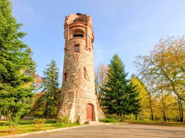Historischer steinerner Bismarckturm in Spremberg, umgeben von grünen und herbstlichen Bäumen unter blauem Himmel. Der zylindrische Turm hat eine rote Ziegelspitze.