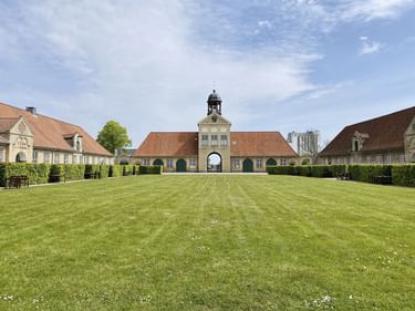 Augustenborg Slot Palastkomplex mit gelben Gebäuden, roten Ziegeldächern und Glockenturm. Gepflegter Rasen im Vordergrund unter blauem Himmel.