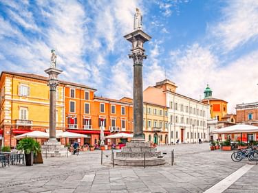 Zwei hohe Steinsäulen mit Statuen stehen auf einem gepflasterten italienischen Platz, umgeben von bunten Gebäuden, Straßencafés und Fahrrädern.