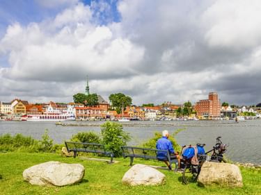 Radfahrer rasten auf Bänken am grünen Ufer in Kappeln. Historische Altstadt mit bunten Häusern und Kirchturm am gegenüberliegenden Hafen.