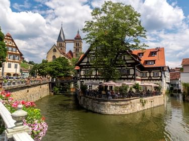 Half-timbered building on stone island in canal in Esslingen old town. Church towers and traditional houses visible under blue sky with clouds.
