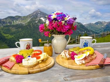 Zwei Holzbrettchen mit österreichischer Brettljause mit Käse, Schinken, Eiern und Gemüse, serviert mit Kaffee und Honig vor Bergkulisse.