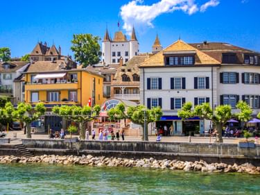 Schloss Nyon mit weißen Türmen erhebt sich über bunten Gebäuden entlang der Seepromenade. Menschen flanieren am Ufer unter blauem Himmel.