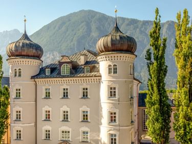 White Lienz Town Hall with two copper onion domes against mountain backdrop. Tall poplar trees frame the baroque-style building under clear blue sky.