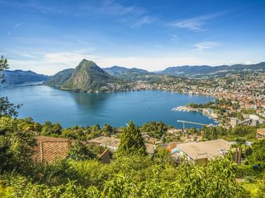 Malerischer Blick auf die Stadt Lugano und den Luganersee mit dem Berg Monte San Salvatore. Blaues Seewasser umgeben von Bergen und Wohngebieten unter klarem Himmel.