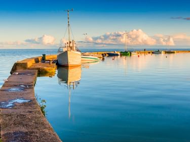 Weißes Fischerboot am Betonpier im Hafen von Nida. Ruhiges Wasser spiegelt Boot und Himmel. Weitere Boote im Hintergrund unter blauem Himmel.