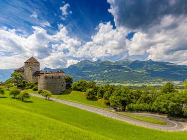 Schloss Vaduz auf einem grünen Hügel mit kurvenreicher Straße, umgeben von Bäumen. Alpine Berge und dramatische Wolken im Hintergrund.