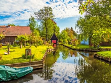 Ruhiger Kanal in Lehde mit Wolkenspiegelung, gesäumt von grünen Rasenflächen und Bäumen. Traditionelle Häuser und ein Bootssteg sind am Wasserweg sichtbar.