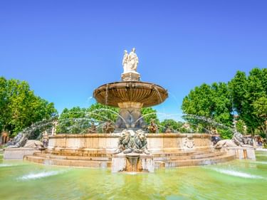 Großer verzierter Brunnen mit mehreren Ebenen und Skulpturen in Aix-en-Provence, umgeben von grünen Bäumen unter blauem Himmel.